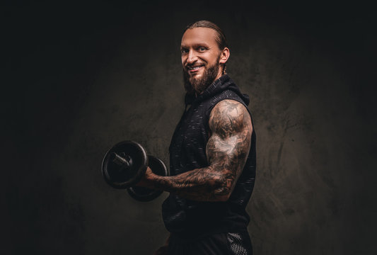Smiling bearded tattooed athlete in a black hoodie posing with a dumbbell in hand. Isolated on dark textured background.
