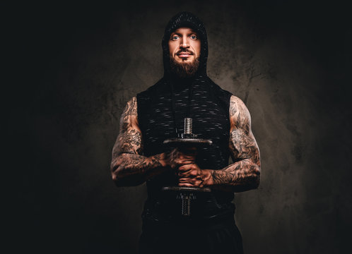 Portrait of a bearded tattooed athlete in a black hoodie posing with a dumbbell in hand.