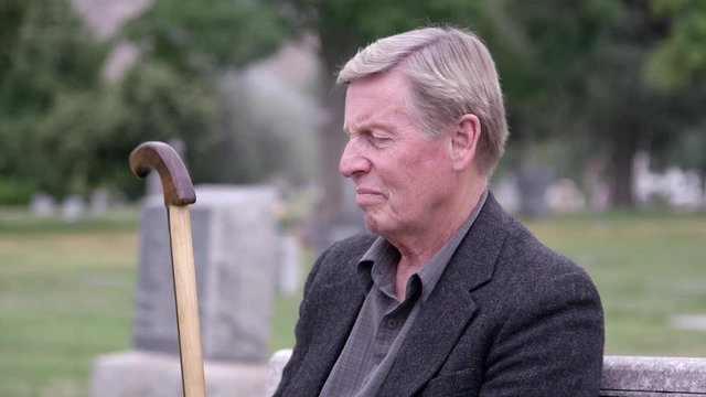 Elderly man sitting at cemetery as he fidgets with his cane being lost in thought.