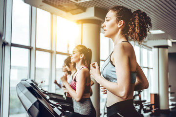 Beautiful young cheerful woman in sportswear running on treadmill at gym with other women