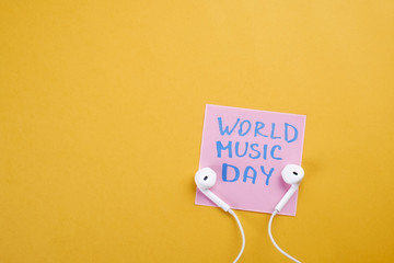 Pair of white earphones and pink sticker paper with World Music Day text written on it by hand. Conceptual composition with earbuds. Bright yellow background, top view, close up, copy space, flat lay.