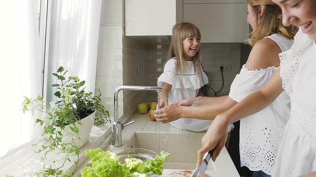 Young caucasian mother with her daughter is having fun splashes each other water in the kitchen and wash the vegetables for ready to cook salad for lunch