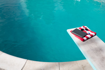 Folded red and white striped beach towel folded on a diving board with a book, sunscreen, and aviator sunglasses on top. Swimming pool in the backyard with folded towel and book, no people.