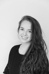 Obraz premium Cute brunette with long hair and black dress looking at camera and smiling with teeth, shoulders turned. White wall background. B&W portrait of cute Caucasian girl with long hair wearing black dress.