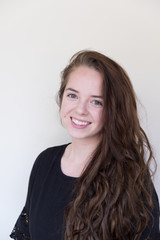 Naklejka premium Cute brunette with long hair and black dress looking at camera and smiling with teeth, shoulders turned. White wall background. Portrait of cute Caucasian girl with long hair wearing black dress.