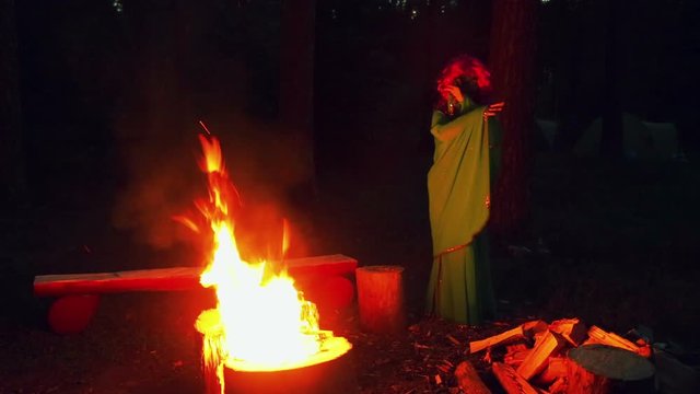 The green witch is warmed by the fire at night in the forest. Seeing someone is covering his face.