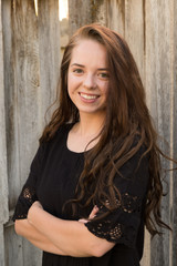 Fototapeta premium Brunette woman posing in front of a rustic old wooden fence wearing a black dress standing with arms crossed. Portrait of a young white woman standing in front of a fence outside in the summer.