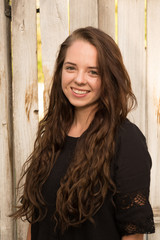 Cute brunette in a black dress standing with arms at her side standing next to a old wooden fence. Young Caucasian girl posing for a head shot in black dress with long brown hair.