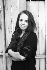 Fototapeta premium Brunette woman posing in front of a rustic old wooden fence wearing a black dress standing with arms folded 3/4 view. Portrait of a young Caucasian woman standing in front of a fence outside, b&w.