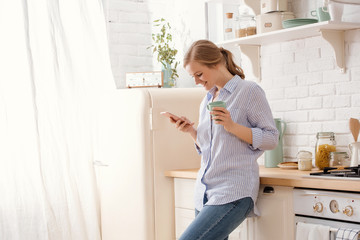 Young woman using smartphone leaning at kitchen table with coffee mug and organizer in a modern home. Smiling woman reading phone message. Brunette happy girl typing a text message.
