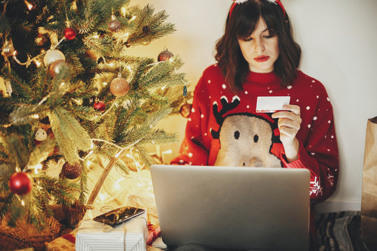 beautiful young woman in reindeer antlers holding credit card, sitting with laptop at golden beautiful christmas tree with lights and presents in festive room. christmas shopping and sales