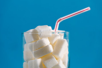 Glass with red stripes straw filled with sugar cubes on blue background. Junk food, unhealthy diet,...
