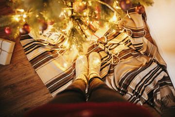 girl legs in stylish warm sock standing with garland lights under christmas tree with presents and gifts. socks on rug in festive room. decor for winter holidays. atmospheric moment