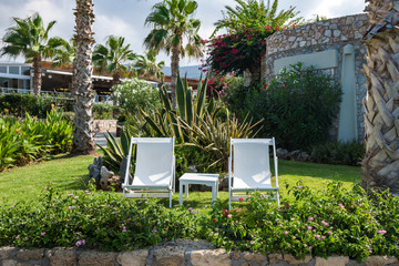 Two empty white tropical deck chairs and a table between standing on a green lawn surrounded by tropical greenery and resort in the background.