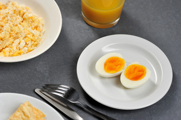 Two halves of hard-boiled egg on a white plate. In the frame of the edge of the plate with scrambled eggs, cutlery, a glass with juice. Gray background. View from above. Close-up. 