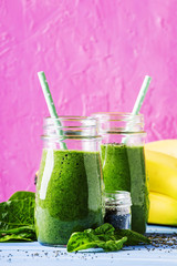 Green smoothies in glass bottles on cool pink blue background with yellow bananas, selective focus
