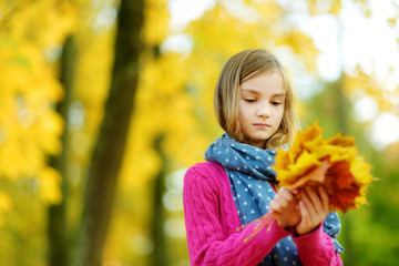 Fototapeta premium Cute little girl having fun on beautiful autumn day. Happy child playing in autumn park. Kid gathering yellow fall foliage.