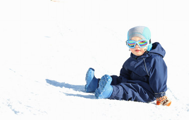 Cute toddler boy in winter suit and ski glusses sits on snow slope on sunny day. Copy space