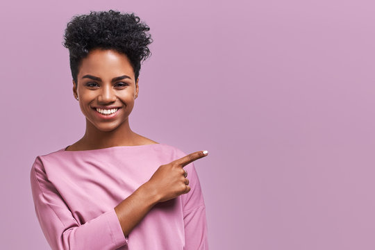 Horizontal portrait of happy dark skinned mixed race female model indicates with fore finger at blank copy space, shows place for your advertisement or promotional text. Lovely African American girl