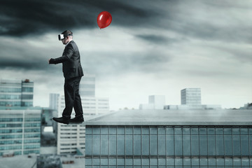 Businessman mit VR Brille und Luftballon in der Hand läuft dem Abgrund eines Wolkenkratzers entlang