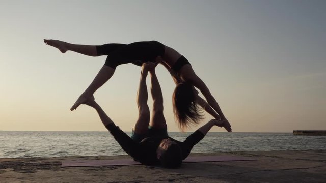 Silhouettes of fit young couple doing acro-yoga at sea beach. Man lying on concrete plates and balancing woman in his feet. Beautiful couple practicing yoga together.