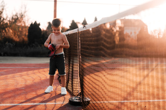 Little boy standing on tennis playground and putting gloves on his hands. Preparing him self for outside training.