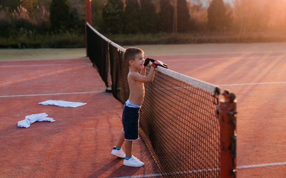 Cute happy little boy standing in tennis playground leaned on tennis net .