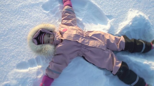 Winter fun snow vacation. Little girl makes a snow angel lying in the snow on a frosty sunny day. Slow Motion