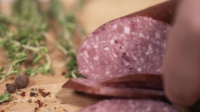 Tasty red meat kielbasa being sliced with metal knife on wooden cutting board next to green herbs at kitchen, close up