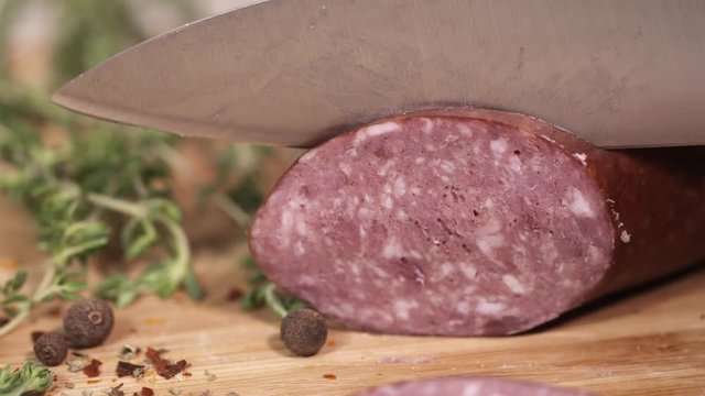 Fresh red meat kielbasa being sliced with metal knife on wooden cutting board next to green herbs at kitchen, close up