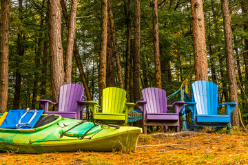 Colorful wooden chairs at rivers edge in the colors of Autumn, with kayak, and brightly colored rope hammock.