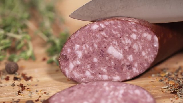 Tasty red meat kielbasa being cutted with metal blade on wooden cutting board next to green herbs at kitchen, close up