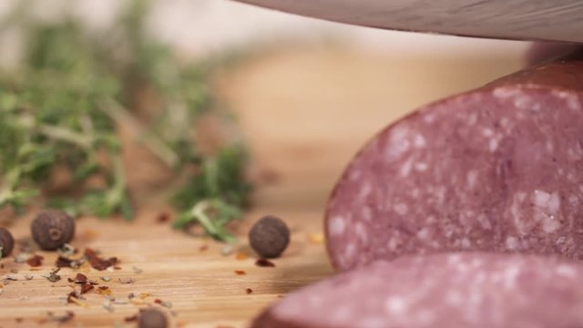 Fresh red meat kielbasa being sliced with metal blade on wooden cutting board next to green herbs at kitchen, close up