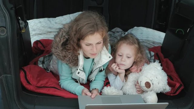 Beautiful young woman and her little daughter are sitting in the open trunk of a car on the river bank of the sea enjoying a laptop