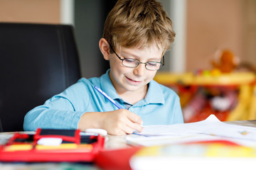 Cute little kid boy with glasses at home making homework, writing letters and doing maths with...