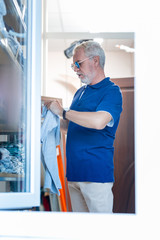 Scrutinizing. Fashionable grey haired man holding T-shirt in hands while looking at it with thoughtful expression on his face