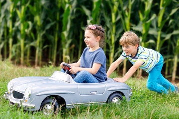 Two happy children playing with big old toy car in summer garden, outdoors. Boy driving car with little girl inside. Laughing and smiling kids. Family, childhood, lifestyle concept..