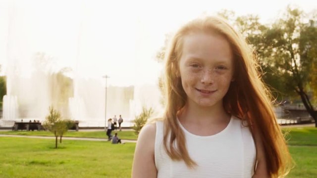 Happy little red-haired girl with freckles walking in sunlight at summer park