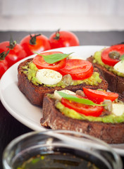 Sandwich toasts with tomatoes cherry, mozzarella, avocado, basil and olive oil. Side view on a dark stone dish.
