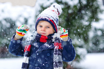 Cute little funny child in colorful winter fashion clothes having fun and playing with snow, outdoors during snowfall. Active outdoors leisure with children. Kid boy and toddler catching snowflakes.