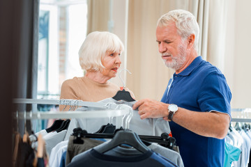 Unsuitable collection. Dissatisfied grey haired man looking down and holding a hanger with T-shirt in hand while his wife standing nearby and cheering him up