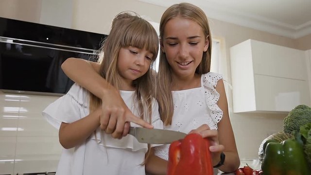 Two teen girls in white dress cook in the kitchen: The girls cuts red peppers to make a salad. Older sister teaches the younger sister to cut paprika. The girls prepare vegetable salad at home in the