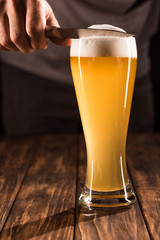 partial view of man taking off beer foam from mug by knife at wooden table
