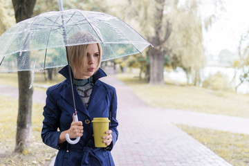 A woman of 30-40 years in a blue raincoat under a transparent umbrella walks in the park and drinks...
