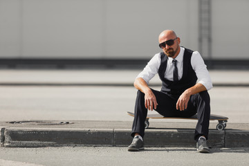 stylish businessman sitting on longboard near modern building