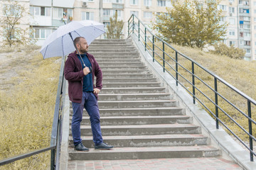 A middle-aged man with a beard on the stairs under an umbrella.