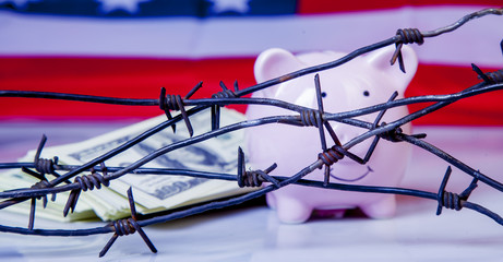 Pink piggy bank and US Dollar money wrapped in barbed wire against United States national flag as...