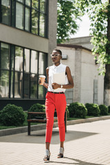 stylish attractive african american businesswoman walking on street with coffee in paper cup and laptop