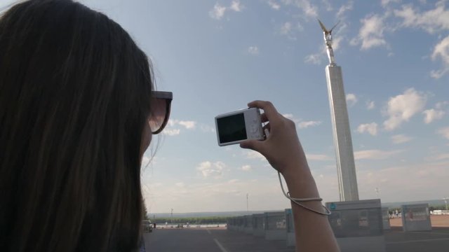 Back view of young white caucasian woman with long dark hair standing on Glory square and taking picture of The Monument of Glory with small white photo camera in summer day in Samara city, Russia