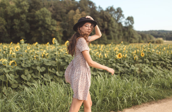 Young brunette girl with a hat and glasses posing and walking in the middle of a countryside of sunflowers